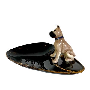 May include: A vintage ashtray featuring a ceramic Boxer dog figurine. The ashtray is oval-shaped, with a glossy black finish and gold trim. The dog is tan and white, wearing a blue collar.