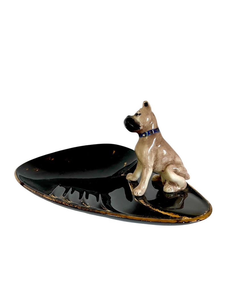 May include: A vintage ashtray featuring a ceramic Boxer dog figurine. The ashtray is oval-shaped, with a glossy black finish and gold trim. The dog is tan and white, wearing a blue collar.