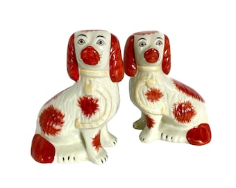 May include: Pair of ceramic dog figurines, each with white and brown speckled fur, sitting upright with their heads tilted slightly to the side. The dogs have red ears and noses, and are wearing yellow collars.