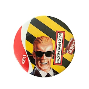 May include: Round button featuring Max Headroom wearing sunglasses, with a yellow and black striped background. The button includes the Coca-Cola logo and the text "MAX HEADROOM".