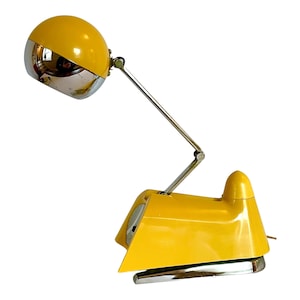 May include: A yellow and chrome desk lamp with a spherical head and adjustable arm. The lamp has a retro design with a bright yellow finish and a chrome base and accents. The lamp is designed to sit on a desk or table.