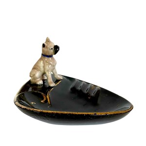 May include: A black ashtray with a gold rim and a ceramic dog figurine. The dog is tan with a black nose and ears, and a blue collar. The ashtray has a leaf-like shape.