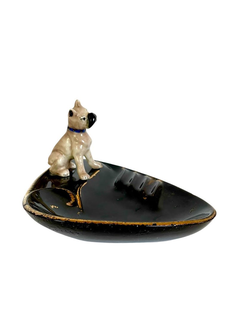 May include: A black ashtray with a gold rim and a ceramic dog figurine. The dog is tan with a black nose and ears, and a blue collar. The ashtray has a leaf-like shape.