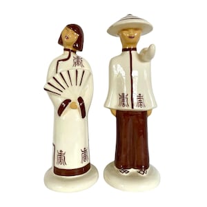 May include: A pair of ceramic figurines depicting a man and woman in traditional Asian clothing. The woman holds a fan and the man wears a conical hat. Both figurines have brown accents and the word "青" written on their clothing.