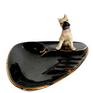 May include: A vintage black ashtray with a gold trim and a ceramic dog figurine. The ashtray has a glossy finish and a unique shape. The dog figurine is tan with a blue collar.