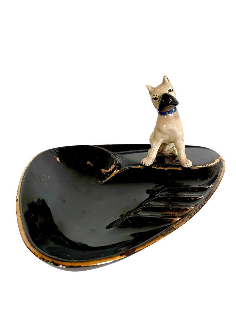 May include: A vintage black ashtray with a gold trim and a ceramic dog figurine. The ashtray has a glossy finish and a unique shape. The dog figurine is tan with a blue collar.