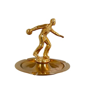 May include: A gold-colored bowling figurine in a throwing pose, mounted on a round, shallow dish. The figure is detailed, with a ball in hand and a focused expression. The dish has a simple, elegant design.