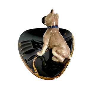 May include: A ceramic ashtray featuring a seated dog figurine. The ashtray is black with gold trim and has a molded design. The dog is light brown with a blue collar. A decorative item.