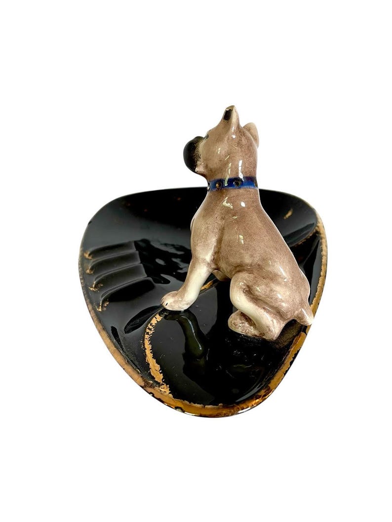 May include: A ceramic ashtray featuring a seated dog figurine. The ashtray is black with gold trim and has a molded design. The dog is light brown with a blue collar. A decorative item.