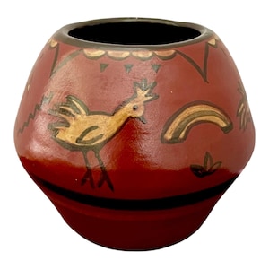 May include: A rounded terracotta-colored ceramic vase featuring a dark brown rim and base. The exterior is decorated with hand-painted illustrations in black and tan, including a stylized bird, a rainbow, and geometric patterns around the top.