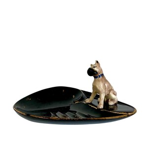May include: A black ashtray with a gold rim and a ceramic Boxer dog figurine. The dog is tan and white with a black snout and a blue collar. The ashtray has a leaf-like shape.