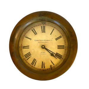 May include: Antique brass clock with a cream-colored face and black Roman numerals. The clock face features the text "Panama Pacific Steamship Co. San Francisco" and "6 Day Patented". The clock has a round shape.