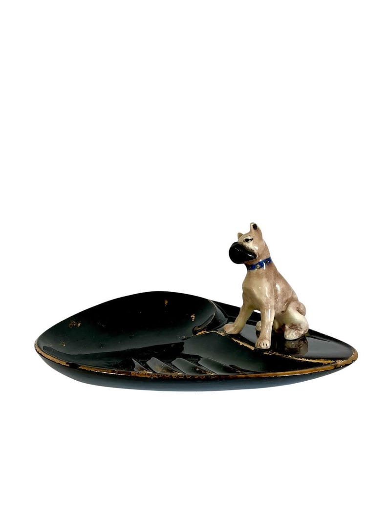 May include: A black ashtray with a gold trim and a ceramic dog figurine. The ashtray has a leaf-like shape with a resting spot for a cigarette. The dog is tan and white with a black nose and a blue collar.