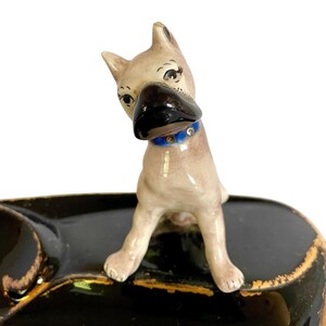 May include: A vintage ceramic dog figurine with a black nose and a blue collar. The dog is perched on a black ashtray with gold trim. The figurine has a light brown coat and is looking to the side.