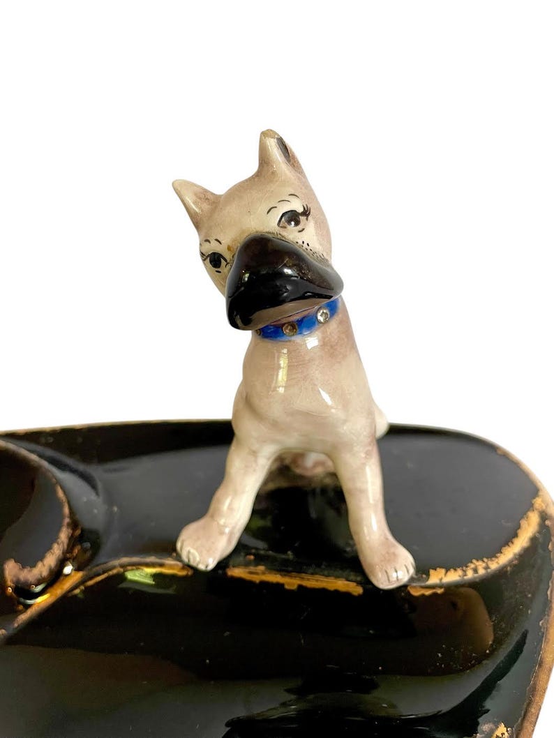May include: A vintage ceramic dog figurine with a black nose and a blue collar. The dog is perched on a black ashtray with gold trim. The figurine has a light brown coat and is looking to the side.