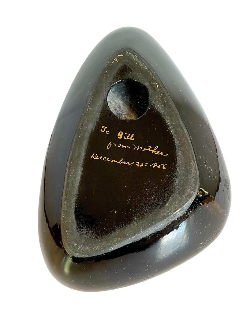 May include: A black, triangular-shaped object with a glossy finish. The underside features a circular opening and handwritten text in gold ink that reads "To Bill from Mother December 25, 1956."
