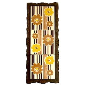May include: A decorative wall hanging featuring three rectangular tiles with a floral design. The tiles have a cream background with vertical brown and white stripes. Brown and yellow flowers are arranged across the tiles, framed by a dark brown wooden border.