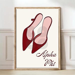 May include: A framed print featuring a pair of red high-heeled shoes with pointed toes and light pink interiors. The words "Alpha Phi" are written in a cursive font below the shoes. The artwork is set against a cream-colored background.