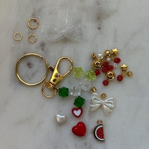 May include: A kit for making a charm bracelet with a variety of beads, charms, and findings. The kit includes gold-coloured jump rings, a lobster clasp, a gold-coloured key ring, clear elastic string, green, red, and white heart charms, a watermelon charm, a white bow charm, green and clear crystal beads, white pearl beads, red and gold beads, and gold star charms.
