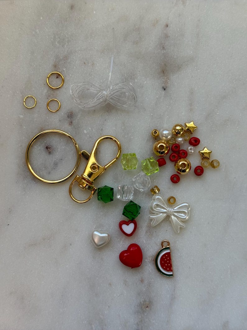 May include: A kit for making a charm bracelet with a variety of beads, charms, and findings. The kit includes gold-coloured jump rings, a lobster clasp, a gold-coloured key ring, clear elastic string, green, red, and white heart charms, a watermelon charm, a white bow charm, green and clear crystal beads, white pearl beads, red and gold beads, and gold star charms.
