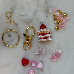 May include: A DIY kit for making a charm bracelet featuring a miniature strawberry cake charm, gold jump rings, a gold lobster clasp, clear elastic string, pink and white heart charms, pink and white pearl beads, gold beads, and clear and pink crystal beads.