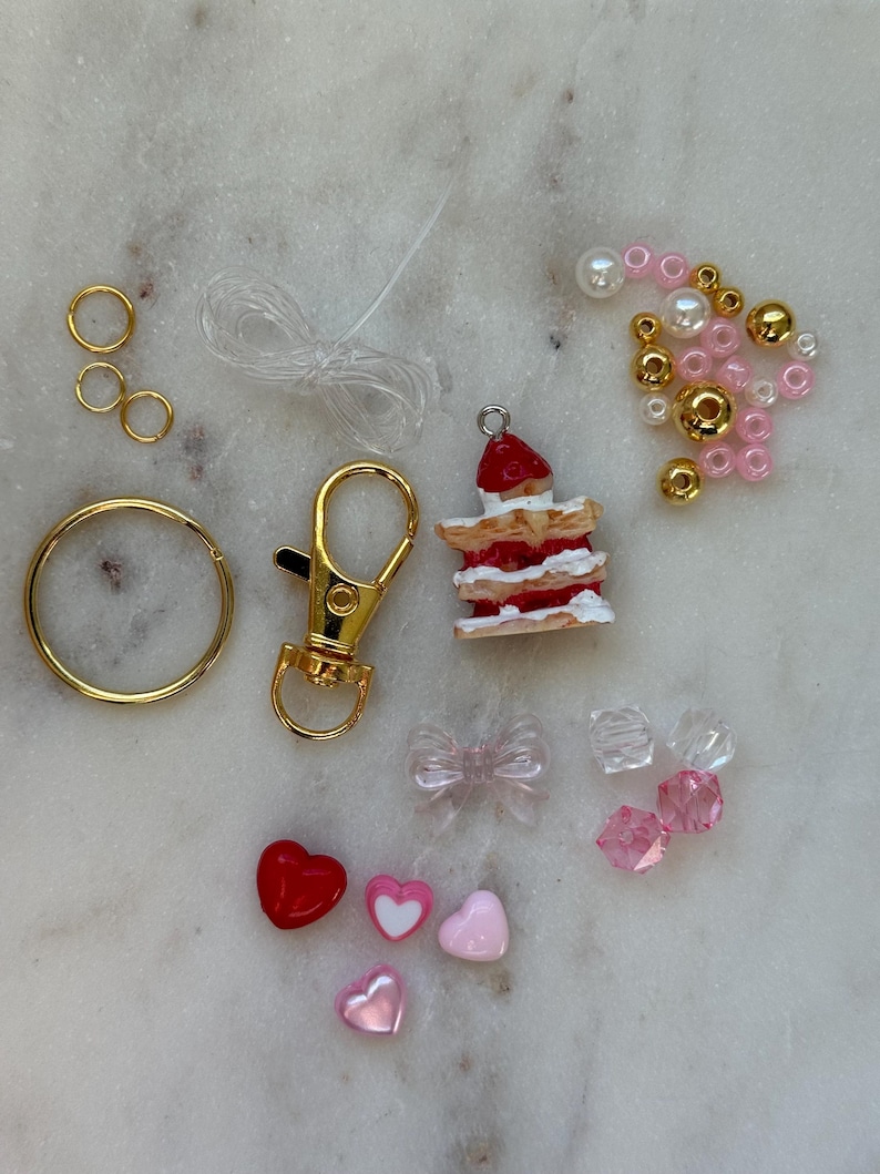 May include: A DIY kit for making a charm bracelet featuring a miniature strawberry cake charm, gold jump rings, a gold lobster clasp, clear elastic string, pink and white heart charms, pink and white pearl beads, gold beads, and clear and pink crystal beads.
