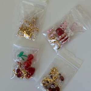 May include: Four clear plastic bags filled with various charms and beads for making keychains. The charms include a star, a cherry, a cake, and a flower. The beads are in shades of red, white, and brown.