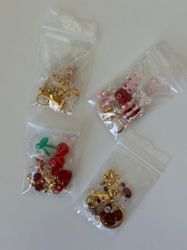 May include: Four clear plastic bags filled with various charms and beads for making keychains. The charms include a star, a cherry, a cake, and a flower. The beads are in shades of red, white, and brown.