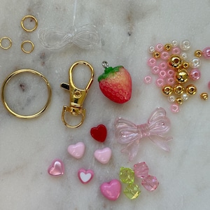 May include: A pink and gold charm kit with a strawberry, hearts, a bow, beads, and a gold key chain.