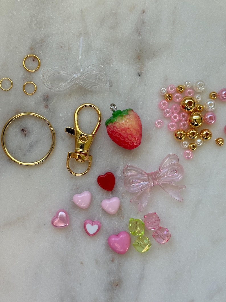 May include: A pink and gold charm kit with a strawberry, hearts, a bow, beads, and a gold key chain.