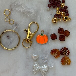May include: A kit for making a fall-themed keychain with a pumpkin charm, gold and maroon beads, clear and brown leaves, a heart, a bow, and a gold key ring.