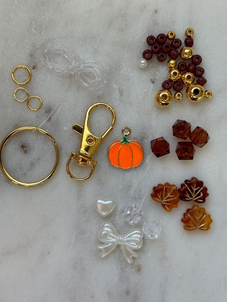May include: A kit for making a fall-themed keychain with a pumpkin charm, gold and maroon beads, clear and brown leaves, a heart, a bow, and a gold key ring.