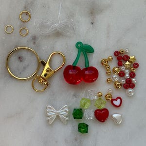 May include: A DIY cherry keychain kit with red and green cherry charms, gold jump rings, a gold lobster clasp, white and red heart charms, green and clear cube beads, white bow charms, and red and white star beads.
