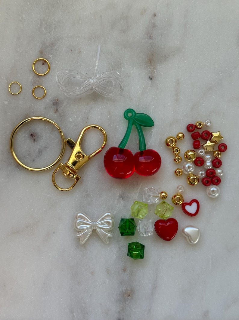 May include: A DIY cherry keychain kit with red and green cherry charms, gold jump rings, a gold lobster clasp, white and red heart charms, green and clear cube beads, white bow charms, and red and white star beads.