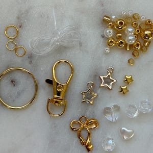 May include: A kit for making a charm bracelet with gold-toned charms, beads, and a clasp. The kit includes a variety of charms, including stars, hearts, and a bow. The beads are a mix of gold and white.