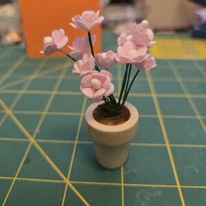 May include: A miniature potted plant with delicate pink flowers and green stems. The flowers have white centers and are arranged in a small, off-white ceramic pot. The pot sits on a green surface with yellow grid lines.