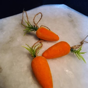 May include: Three orange carrot ornaments with green leafy tops and twine hangers. The carrots are arranged on a white surface, perfect for Easter or spring decor.