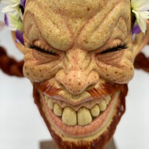 Puede incluir: Un primer plano de una cara esculpida con una sonrisa traviesa. La cara est&aacute; hecha de un material marr&oacute;n claro y tiene una barba y un bigote rojos. La cara est&aacute; adornada con una corona de flores blancas con acentos morados.