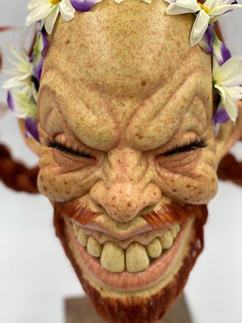 Puede incluir: Un primer plano de una cara esculpida con una sonrisa traviesa. La cara est&aacute; hecha de un material marr&oacute;n claro y tiene una barba y un bigote rojos. La cara est&aacute; adornada con una corona de flores blancas con acentos morados.