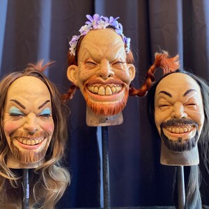 May include: Three realistic-looking, hand-crafted, smiling human heads with different hairstyles and facial features. The heads are mounted on black stands.