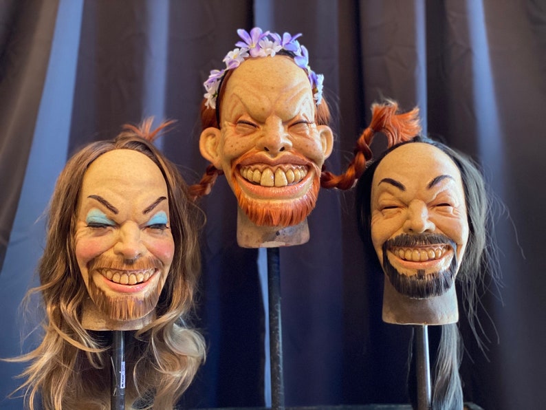 May include: Three realistic-looking, hand-crafted, smiling human heads with different hairstyles and facial features. The heads are mounted on black stands.