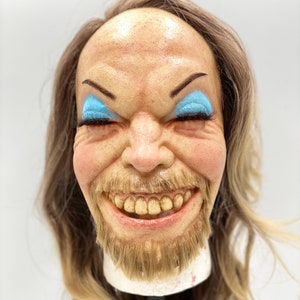 May include: A close-up of a smiling face mask with a long blonde wig. The mask has blue eyeshadow and a brown beard. The mask is on a stand.