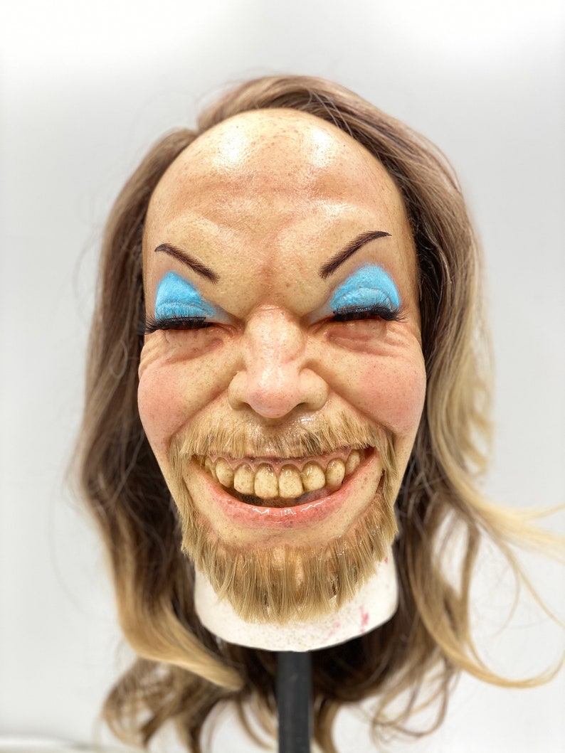 May include: A close-up of a smiling face mask with a long blonde wig. The mask has blue eyeshadow and a brown beard. The mask is on a stand.