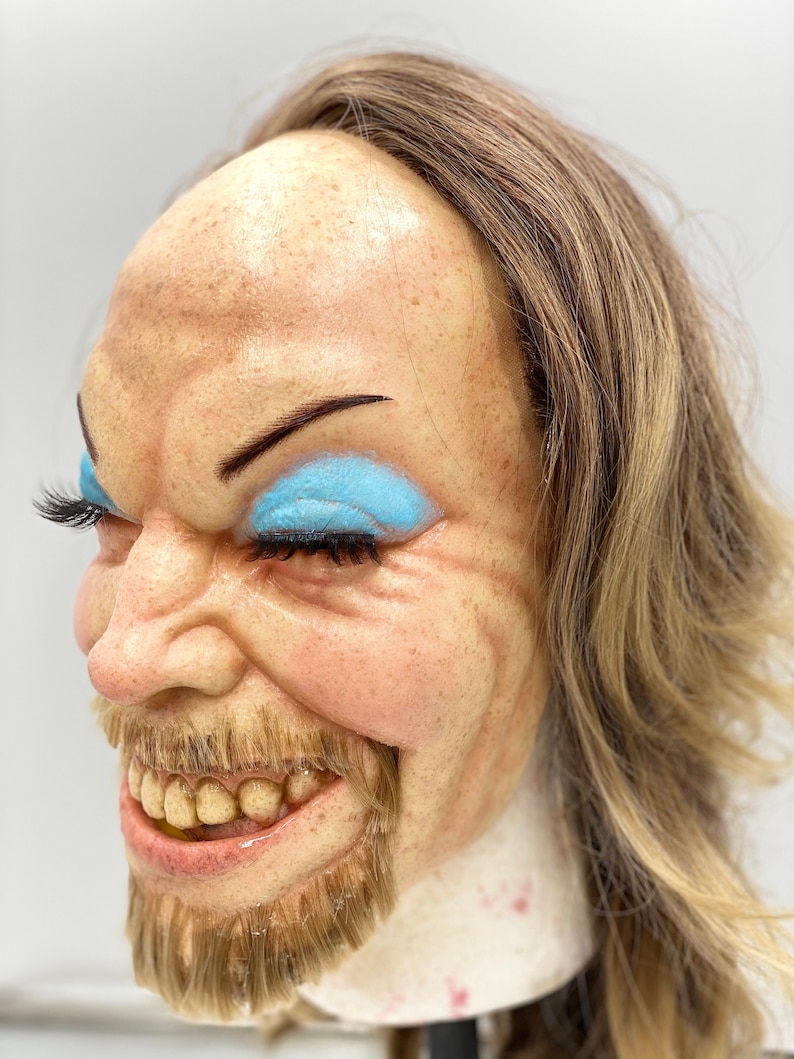 May include: A close-up of a realistic-looking mask with a light brown wig, a wide smile, and blue eyeshadow. The mask has a painted-on beard and mustache.