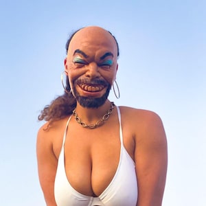 May include: A person wearing a white bikini top with a bald head and a smiling face mask. The mask has a brown complexion, blue eyeshadow, and a wide, toothy grin.
