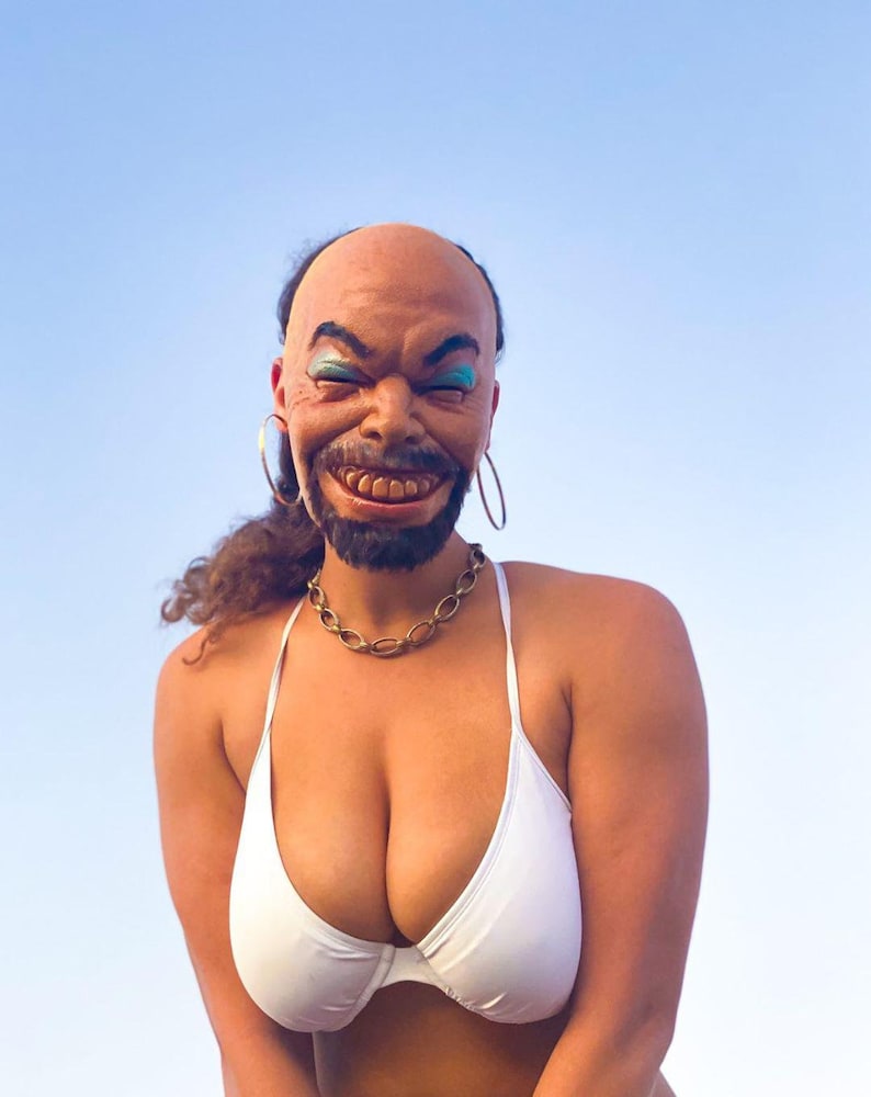 May include: A person wearing a white bikini top with a bald head and a smiling face mask. The mask has a brown complexion, blue eyeshadow, and a wide, toothy grin.