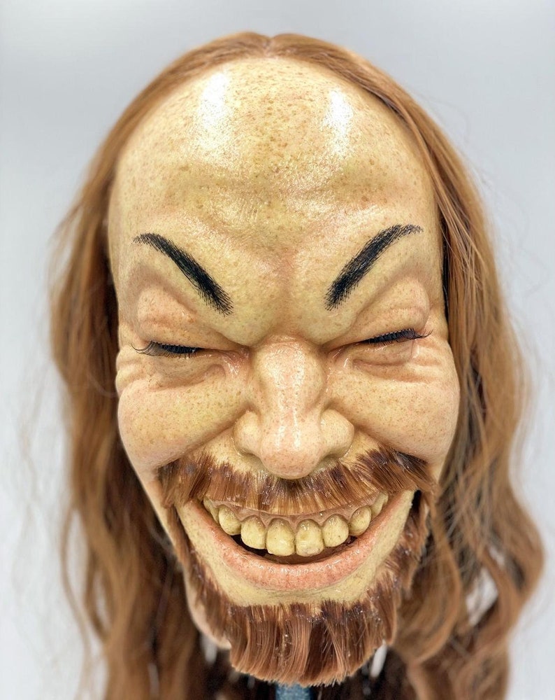 May include: A close-up of a realistic-looking, hand-crafted,  creepy, smiling,  human head with long, brown hair. The head has a painted,  yellowish-tan complexion,  a wide, toothy grin, and a brown beard.