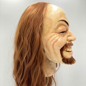 May include: A realistic-looking, hand-painted, sculpted mask of a man with long, wavy, auburn hair and a beard. The mask is smiling with a mischievous expression.