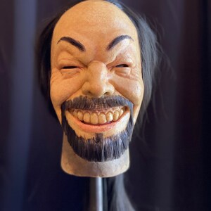 May include: A realistic-looking, hand-painted,  creepy, smiling,  human head mask with a long black wig. The mask has a wide, toothy grin and a dark beard.