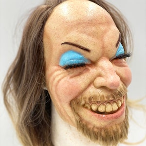 May include: A realistic, hand-painted, sculpted face mask with long brown hair, a beard, and blue eyeshadow. The mask is smiling with its mouth open, showing teeth.